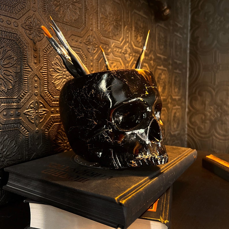 May include: A black skull-shaped ceramic container with a set of paintbrushes inside. The container is sitting on top of a black book with gold lettering.