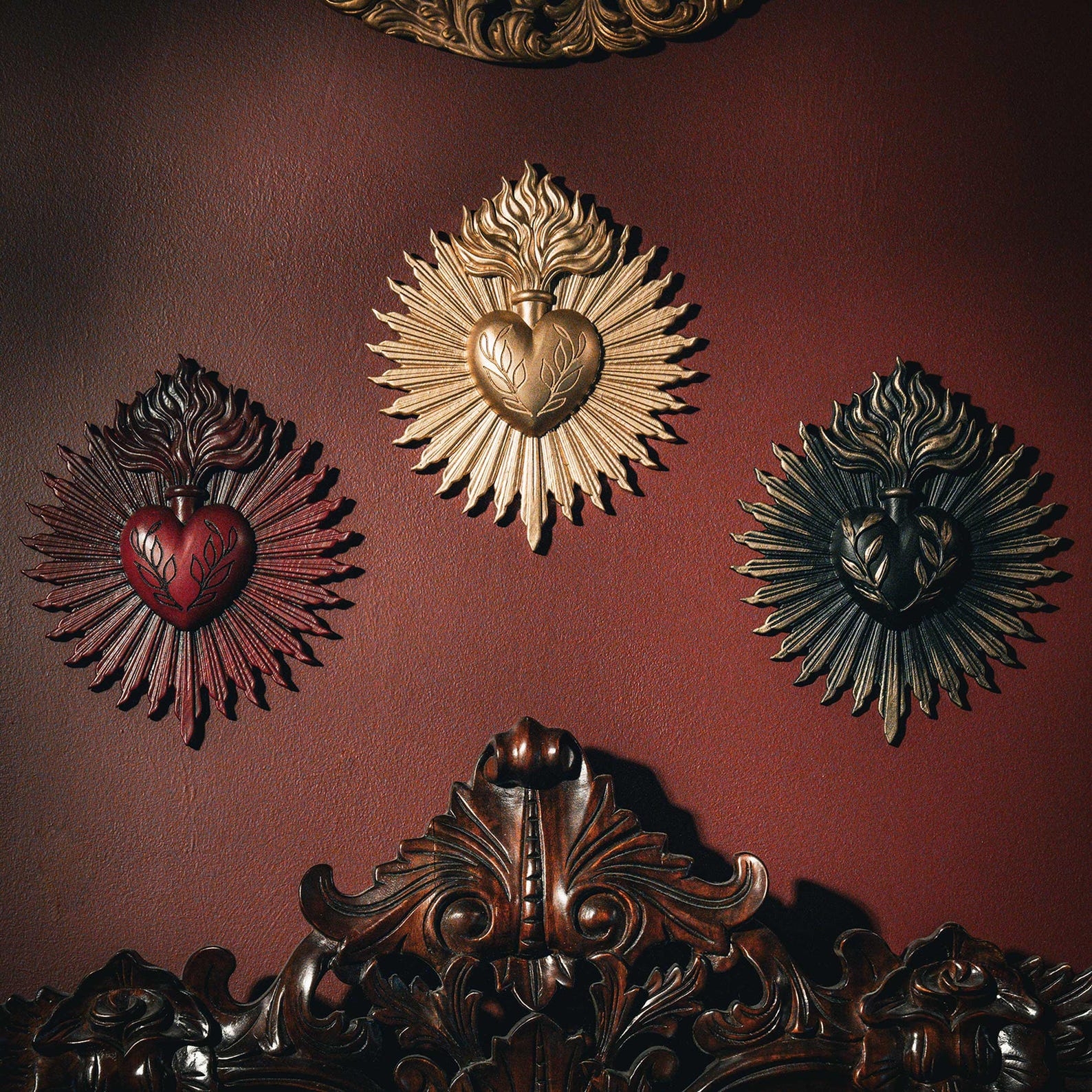 Three sacred heart wall plaques in red, gold, and black with radiant flame designs on a deep red wall above an ornately carved wooden headboard