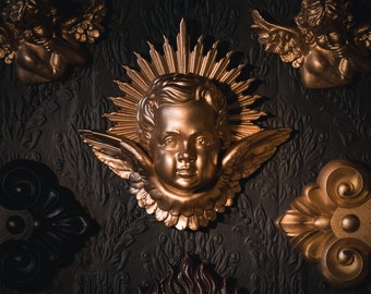 Puede incluir: Una figurita de querubín de oro con alas y halo, rodeada de adornos dorados ornamentados sobre un fondo oscuro.