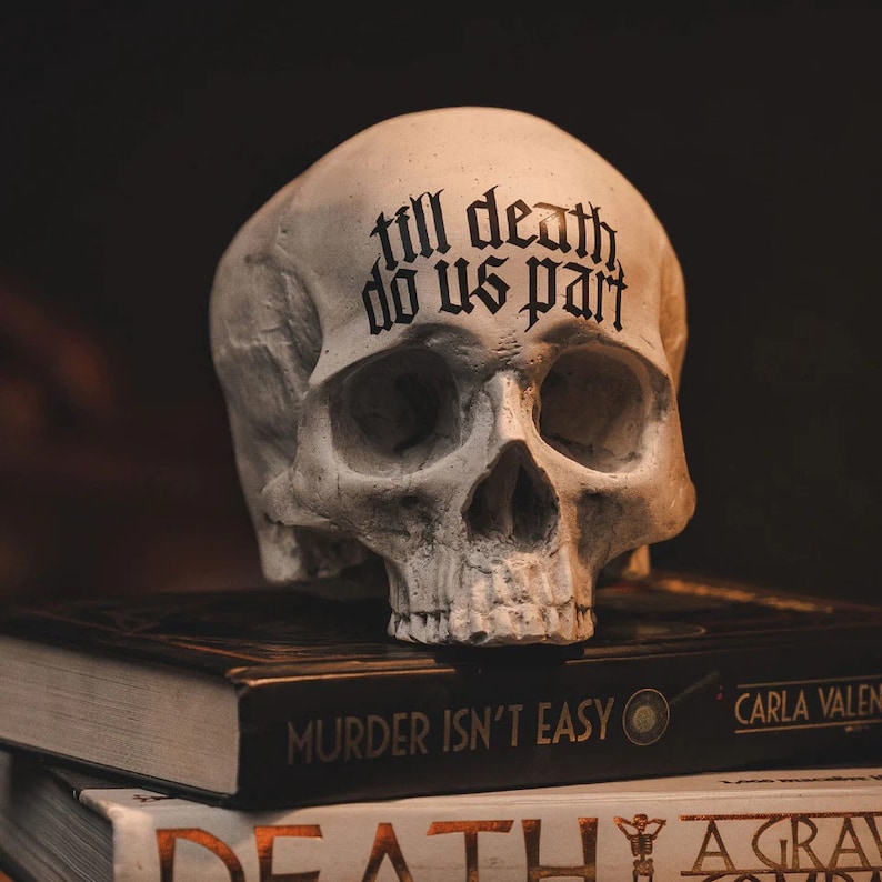 May include: A white skull with the words "till death do us part" written on the forehead, sitting on top of two books. The top book has the title "Murder Isn't Easy" and the bottom book has the title "Death Agrave Concern".