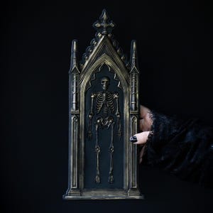 May include: A black and gold gothic-style wall hanging featuring a skeleton figure. The decorative piece has an arched top and intricate detailing, with a dark background. The skeleton is in a standing position.
