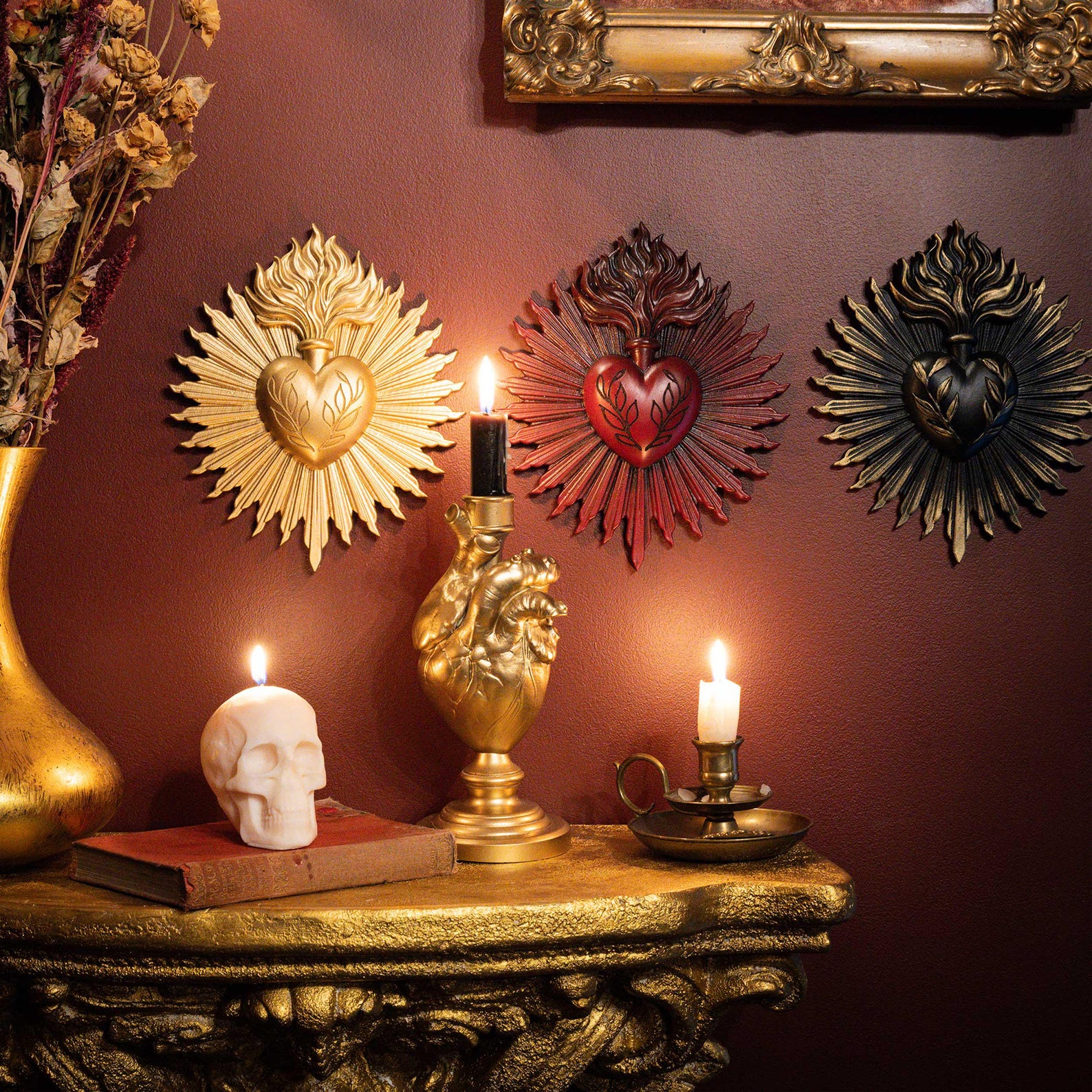 Three sacred heart wall plaques in gold, red, and black above a gold altar table with lit candles, a heart-shaped candlestick, and a skull candle