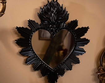 Könnte beinhalten: Ein schwarzer herzförmiger Wandspiegel mit einem dekorativen Rahmen aus strahlenförmigen Sonnenstrahlen und einem skulpturalen Flammenmotiv an der Oberseite. Die Spiegelfläche ist reflektierend und glatt, umgeben von einem dunklen, strukturierten Rand.