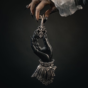 May include: A dark metallic sculpture of a hand holding an ornate key. The hand features a detailed wrist cuff with a bow and lace-like texture. The key has a decorative bow-shaped head and a vertical shaft. The finish is a weathered bronze tone.