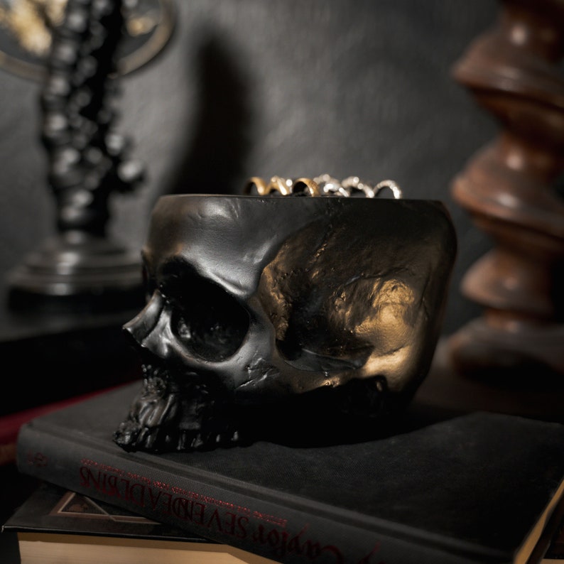 May include: Black skull-shaped ring holder with gold accents. The holder is sitting on a black book with the title "Carver Seven Deadly Sins".