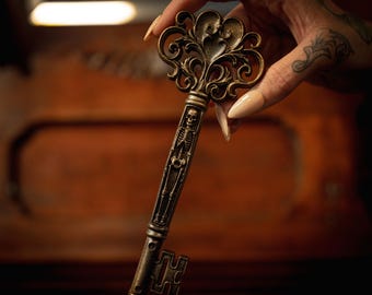 Op de afbeelding: Een antieke messing sleutel met een sierlijke, gekrulde kop en een gedetailleerde skeletfiguur in reliëf langs de schacht. De sleutel heeft een donkere, verweerde afwerking en een rechthoekige baard, met fijn gedetailleerd metaalwerk.