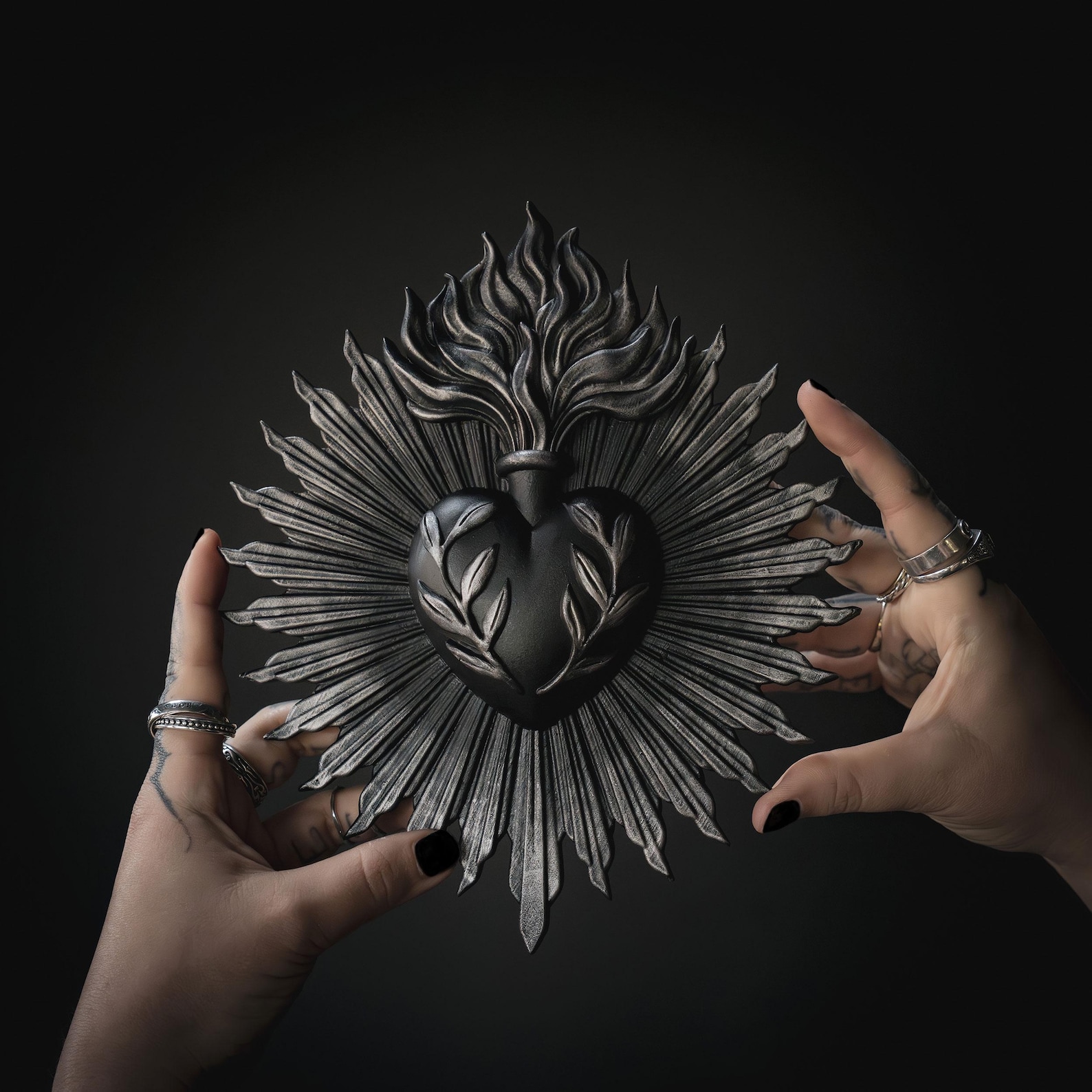 Hands with black nail polish and silver rings holding a black and silver sacred heart wall plaque with flame and leaf detailing against a dark background