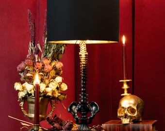 May include: A table lamp featuring a black cylindrical lampshade and a base sculpted to resemble a human spinal column and pelvis. The structure is finished in a dark, matte black tone, providing a stark contrast to the light emitted from within.
