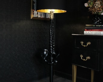 May include: A black floor lamp with a black lampshade and a gold trim. The lamp is shaped like a human spine and has a gold base. The lamp is in a room with black walls and a black dresser with gold handles.