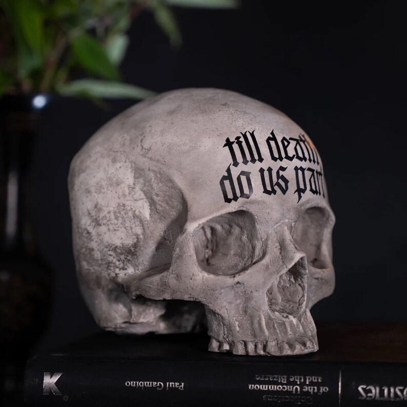May include: A gray human skull with the words "till death do us part" written in black gothic font across the forehead.