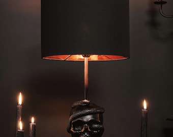 May include: A black table lamp with a skull base and a black lampshade. The skull has a snake wrapped around it and a copper-colored interior. The lamp is surrounded by lit black candles, creating a gothic or Halloween theme.