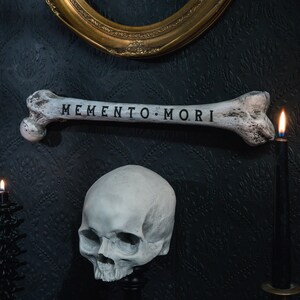 Op de afbeelding: Een witte schedel met een bot waarop de woorden "Memento Mori" staan, op een zwarte achtergrond. Het bot is boven de schedel geplaatst en er zijn twee kaarsen aan weerszijden van de schedel.