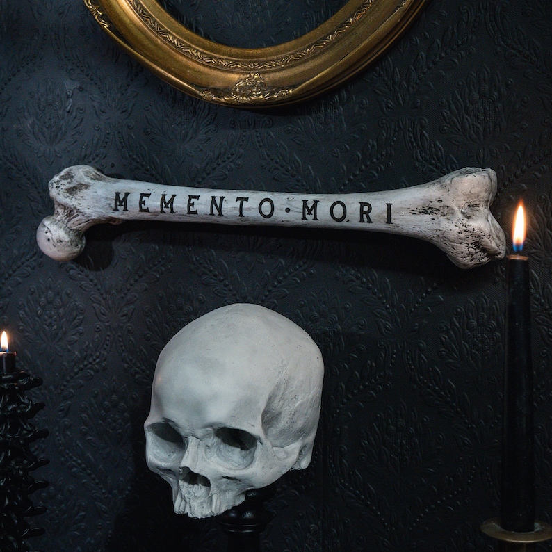 Op de afbeelding: Een witte schedel met een bot waarop de woorden "Memento Mori" staan, op een zwarte achtergrond. Het bot is boven de schedel geplaatst en er zijn twee kaarsen aan weerszijden van de schedel.
