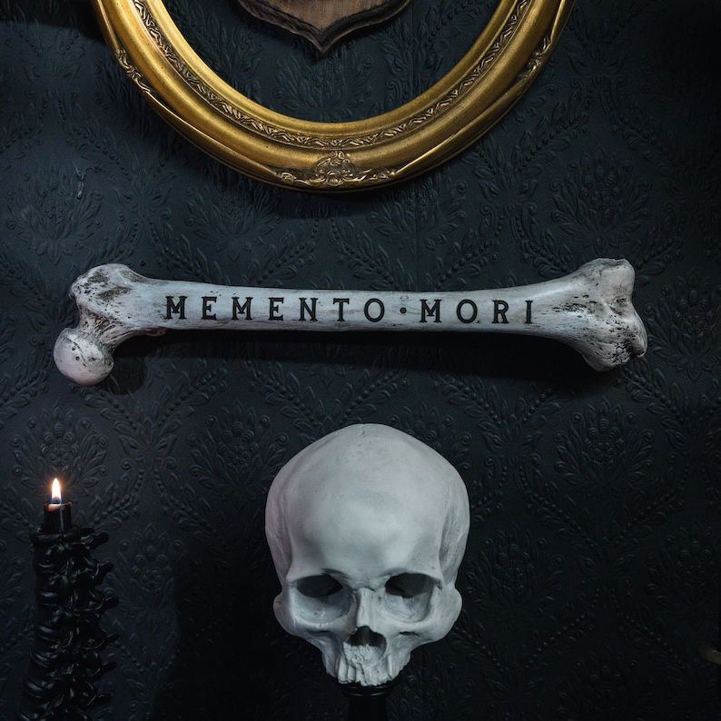 Op de afbeelding: Een witte schedel met een bot waarop "Memento Mori" staat, tegen een donkere achtergrond.