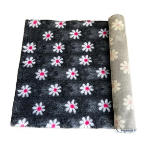 Puede incluir: Una alfombra gris oscuro enrollada con un patrón repetido de margaritas blancas con centros rosas. Se ve la parte inferior de la alfombra, que muestra un diseño floral gris y blanco. La alfombra está sobre un fondo blanco.