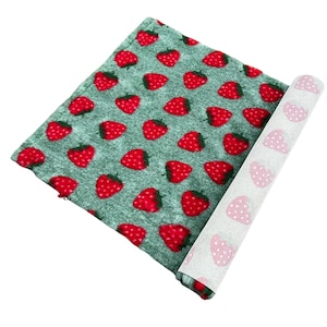 Puede incluir: Una tela verde enrollada con un patrón repetido de fresas rojas. La parte inferior es blanca con un patrón de corazones rojos. La tela es probablemente un artículo decorativo.