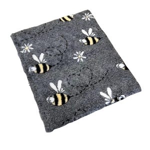 Puede incluir: Una manta de forro polar gris con un patrón de abejas y flores. Las abejas son amarillas y negras, y las flores son blancas.