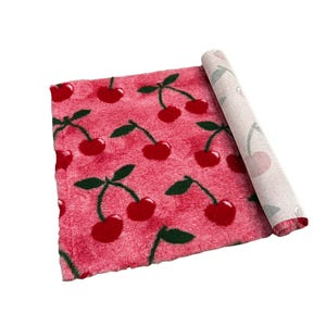 Puede incluir: Una alfombra rosa enrollada con un patrón repetido de cerezas rojas y tallos verdes. La parte inferior de la alfombra es visible, con un color más claro y un diseño de cerezas. La alfombra está hecha de un material suave y afelpado.