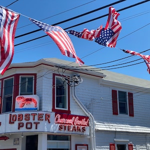 May include: A white building with red trim and a sign that says "Lobster Pot" and "Charcoal Broiled Steaks". There are American flags hanging from the building.