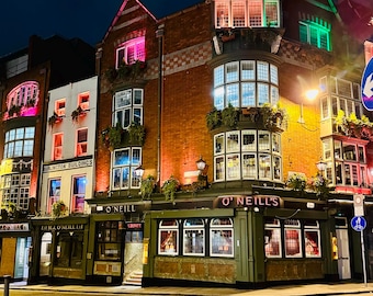 May include: Nighttime exterior shot of O'Neill's pub, a multi-story building with brick and painted facades. The building is illuminated with warm and colorful lights, including green, pink, and orange. The street is empty, and the sky is dark.