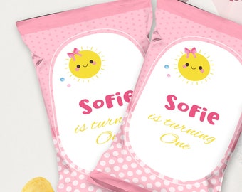 May include: Two pink chip bags with white polka dots and a yellow sun with a pink bow. The text on the bags reads "Sofie is turning One".