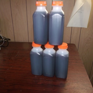 Pu&ograve; includere: Sei bottiglie di plastica trasparente piene di un liquido scuro, impilate a forma di piramide. Le bottiglie hanno corpi bianchi e tappi arancioni. Le bottiglie sono su una superficie di legno marrone scuro.