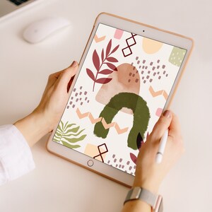 May include: A digital tablet displaying an abstract design with botanical and geometric shapes in shades of green, brown, pink, and peach. The tablet is held by two hands, with a stylus in one hand.