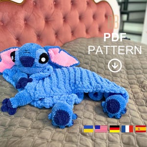 Könnte beinhalten: Blaues Häkeltier in Form des Stitch-Charakters mit rosa Ohren. Das Spielzeug liegt auf einem braunen gesteppten Bettüberwurf. Der Text "PDF PATTERN" ist in Weiß auf schwarzem Hintergrund über dem Spielzeug geschrieben. Ein nach unten zeigender Pfeil befindet sich unter dem Text.
