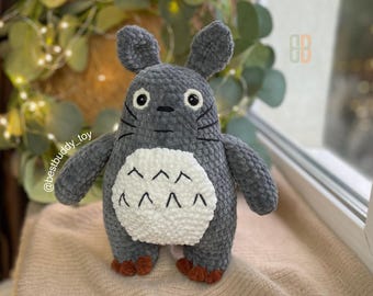 May include: A gray crocheted plush toy resembling a character from the animated film "My Neighbor Totoro". The toy has large, round eyes, a white belly with a pattern of small, white triangles, and a fluffy texture.