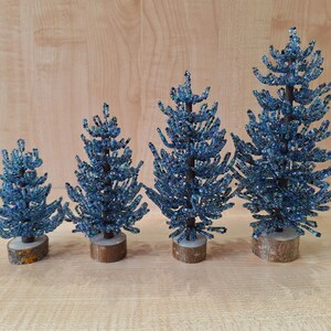 K&ouml;nnte beinhalten: Vier blaue, perlenbesetzte Weihnachtsb&auml;ume in verschiedenen Gr&ouml;&szlig;en auf einem Holzgrund.