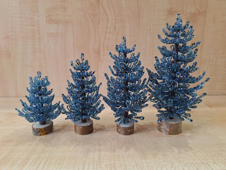 K&ouml;nnte beinhalten: Vier blaue, perlenbesetzte Weihnachtsb&auml;ume in verschiedenen Gr&ouml;&szlig;en auf einem Holzgrund.
