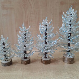 K&ouml;nnte beinhalten: Vier wei&szlig;e Perlen-Weihnachtsb&auml;ume in verschiedenen Gr&ouml;&szlig;en auf einer Holzoberfl&auml;che. Die B&auml;ume sind mit blauen Perlen verziert und haben braune Holzsockel.