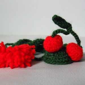 May include: A crocheted cherry garland with two red cherries and green leaves. The garland is made of yarn and is a handmade craft.