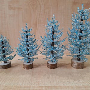 K&ouml;nnte beinhalten: Vier blaue und wei&szlig;e Perlen-Weihnachtsb&auml;ume in verschiedenen Gr&ouml;&szlig;en auf einer Holzoberfl&auml;che.