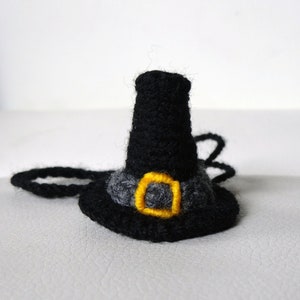 May include: A black felt witch hat with a gray brim and a yellow buckle. The hat is attached to a black cord.
