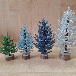 K&ouml;nnte beinhalten: Vier Perlen-Weihnachtsb&auml;ume, ein gr&uuml;ner, zwei blaue und ein wei&szlig;er, auf einer Holzoberfl&auml;che.