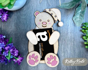 May include: Wooden bear figurine holding an Amazon gift card. The bear is wearing a blue and white sleep cap. The bear has pink paw prints on its feet. The gift card is black with the Amazon logo and the text "amazon.com gift card".