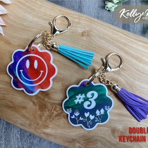 May include: Two double-sided keychains with gold chains and tassels. One keychain features a colorful swirl design with a white smiley face. The other keychain features a colorful swirl design with a white number "3" and white floral accents. The text "DOUBLE SIDED KEYCHAIN DESIGN" is visible at the bottom of the image.