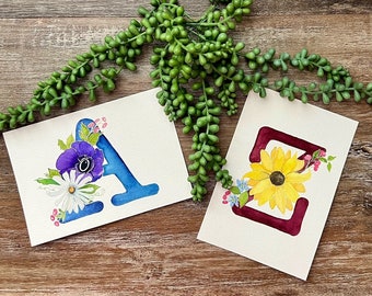 May include: Two watercolor paintings of letters with floral designs. The letter A is blue with a purple and white flower design. The letter Z is maroon with a sunflower design.
