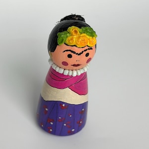 May include: A hand-painted wooden peg doll featuring black hair with a yellow floral crown, a white beaded necklace, a pink shawl over a cream-colored bodice, and a purple skirt with small red and white floral patterns, painted with a distinct unibrow.