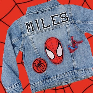 May include: A light blue denim jacket with the name "MILES" in white block letters on the back. The jacket features three Spider-Man patches: a large mask, a small spider symbol, and a figure in a web-slinging pose.