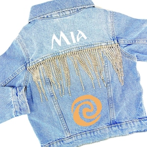 May include: A light blue denim jacket with a gold fringe and rhinestones. The jacket has a white "MIA" written on the back and a brown swirl design.