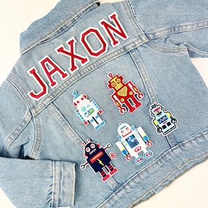 May include: A blue denim jacket with the name "Jaxon" embroidered on the back in red letters. The jacket is decorated with five colorful robot patches.