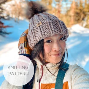 May include: A close-up of a woman wearing a light pink knitted beanie with a fluffy pom-pom on top. The beanie has a ribbed texture and a small leather tag with a logo. The woman is smiling and looking at the camera. The background is blurred and shows a snowy landscape.