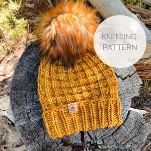 May include: A knitted beanie hat in a mustard yellow color with a fluffy brown pom pom on top. The hat has a ribbed brim and a textured pattern. The hat is sitting on a piece of wood with the text "Knitting Pattern" on a white circle.
