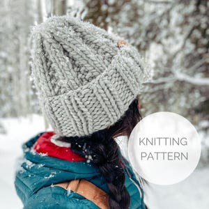 Puede incluir: Un gorro de punto gris con un patrón de punto de cable. El gorro lo lleva una persona con el pelo largo y oscuro. El texto "KNITTING PATTERN" es visible en la imagen.