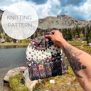 Peut inclure: Un bonnet tricoté coloré avec une bande blanche et une étiquette en cuir marron qui dit "Knit One, Purl Two". Le bonnet est tenu par une main devant un fond de montagne et de lac.
