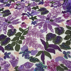 May include: Close-up of a fabric with a floral design. The fabric features a variety of flowers and leaves in shades of purple, pink, and green. The flowers are embellished with sequins, adding a shimmering effect to the design.