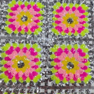 May include: Close-up of a fabric with a repeating floral pattern. The flowers are composed of pink, yellow, and lime green sequins, set against a silver sequined background. The design is symmetrical and eye-catching.