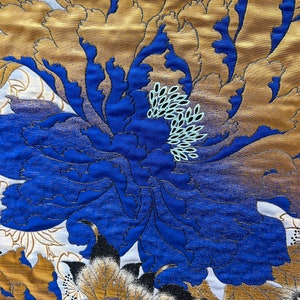May include: Close-up of a fabric with a large floral design. The dominant colors are gold, blue, and white. The central flower is a vibrant blue with gold accents, surrounded by gold and white petals. The fabric has a textured appearance.
